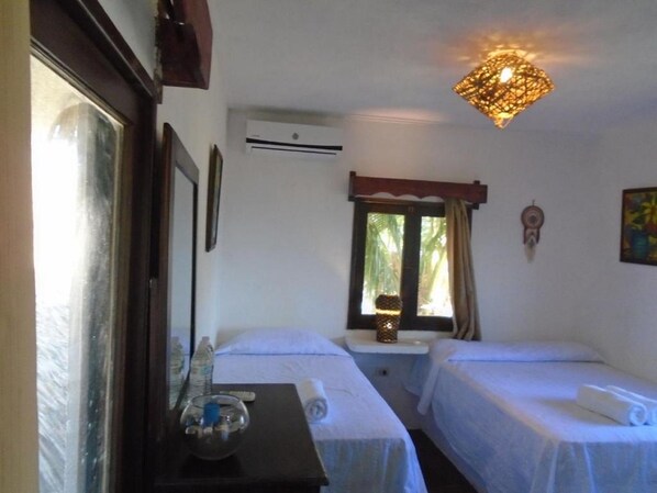 A hotel room with two single beds, a small table with a bottle and a glass, a mirror, a window with a view of greenery, and a decorative wall hanging.