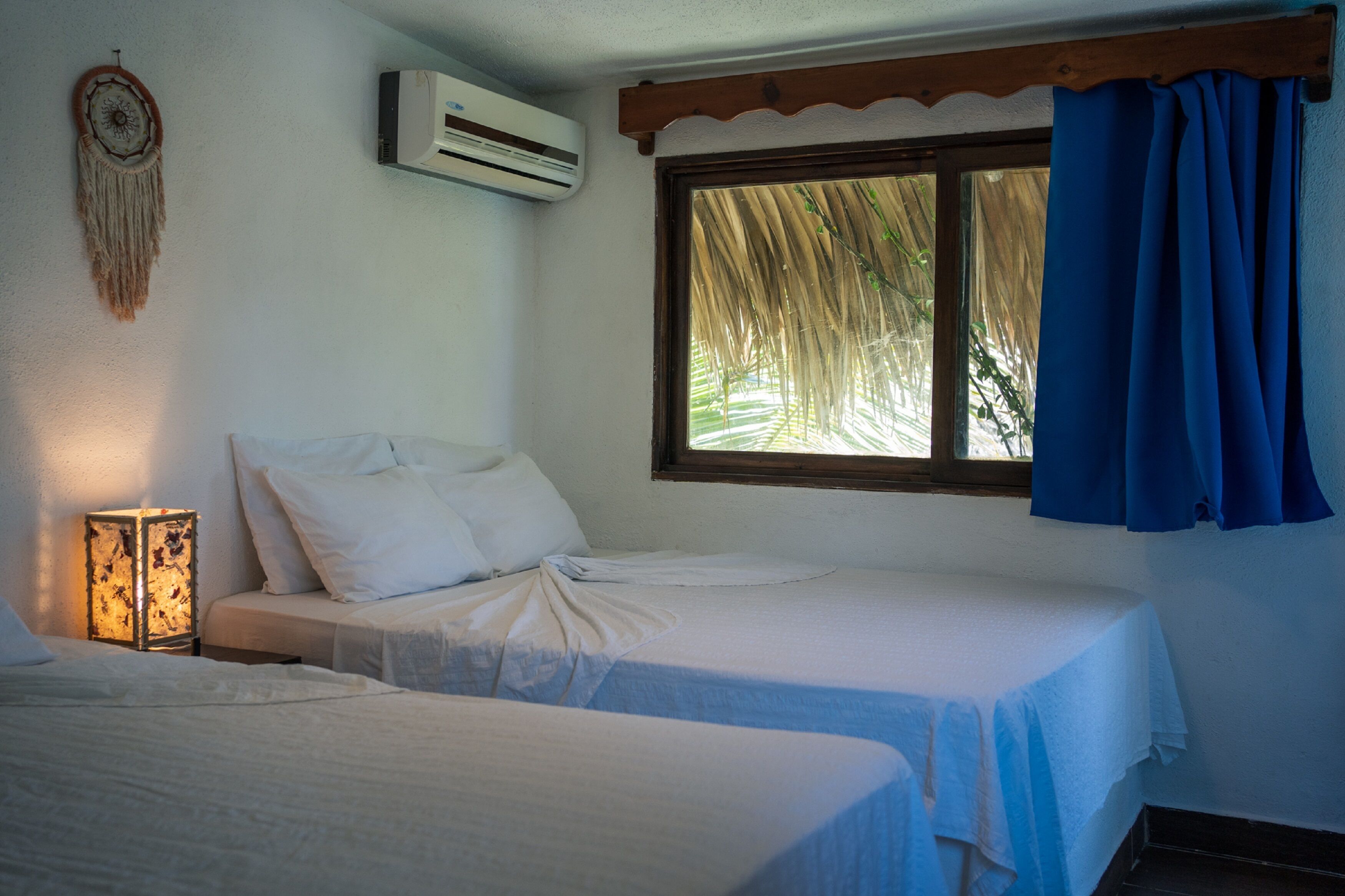Een kamer met twee bedden, een wandairconditioner, een raam met blauwe gordijnen en een decoratieve wandhanger.