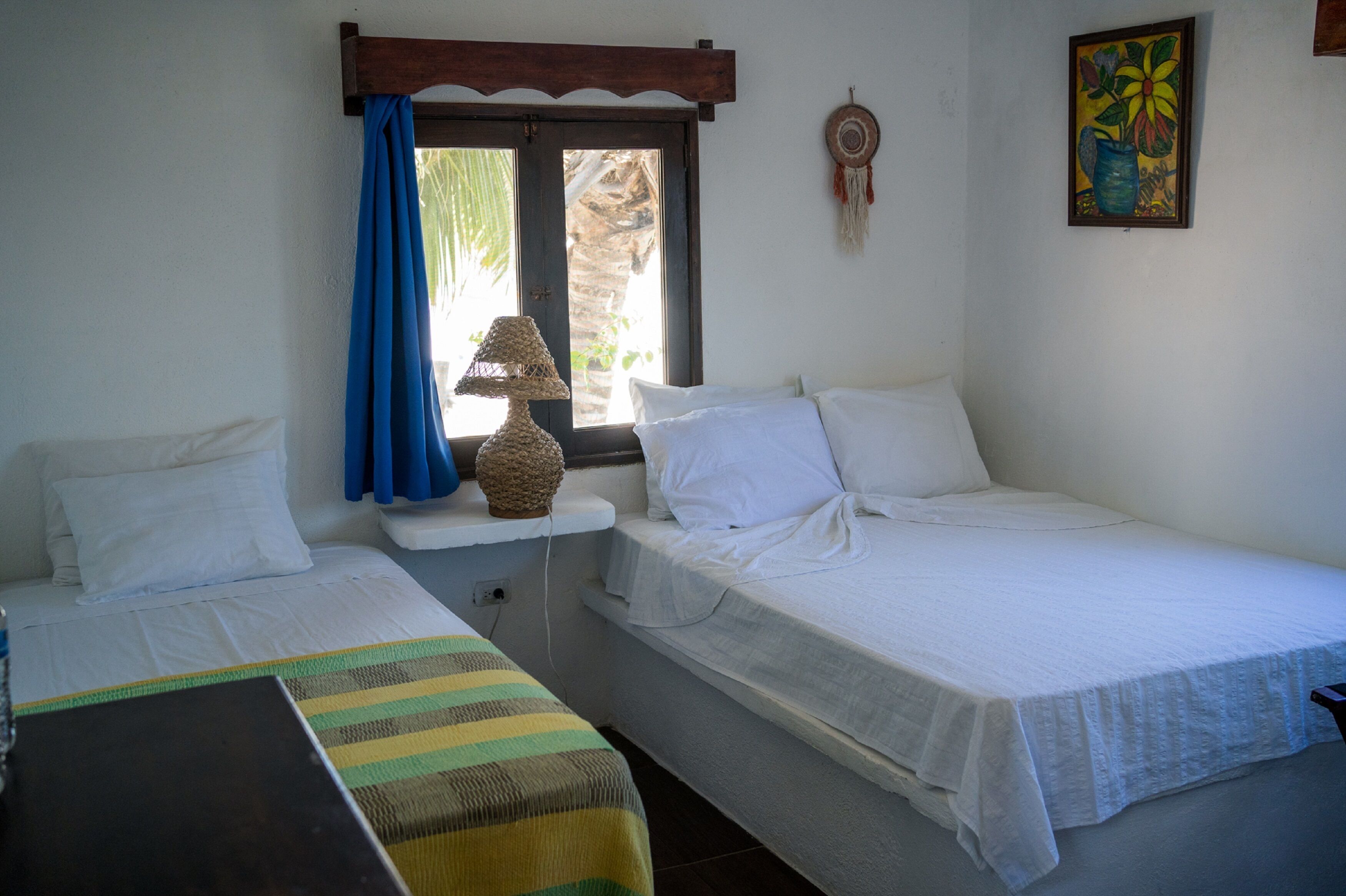 A room with two single beds, a small table with a lamp, and a window with blue curtains.
