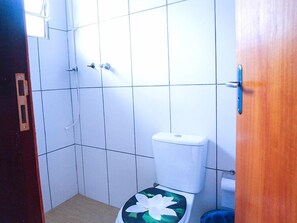 A small bathroom with white tiles, a toilet with a floral seat cover, and a wooden door. - Pousada Montanhas do Bem TeVi (Caraguatatuba)