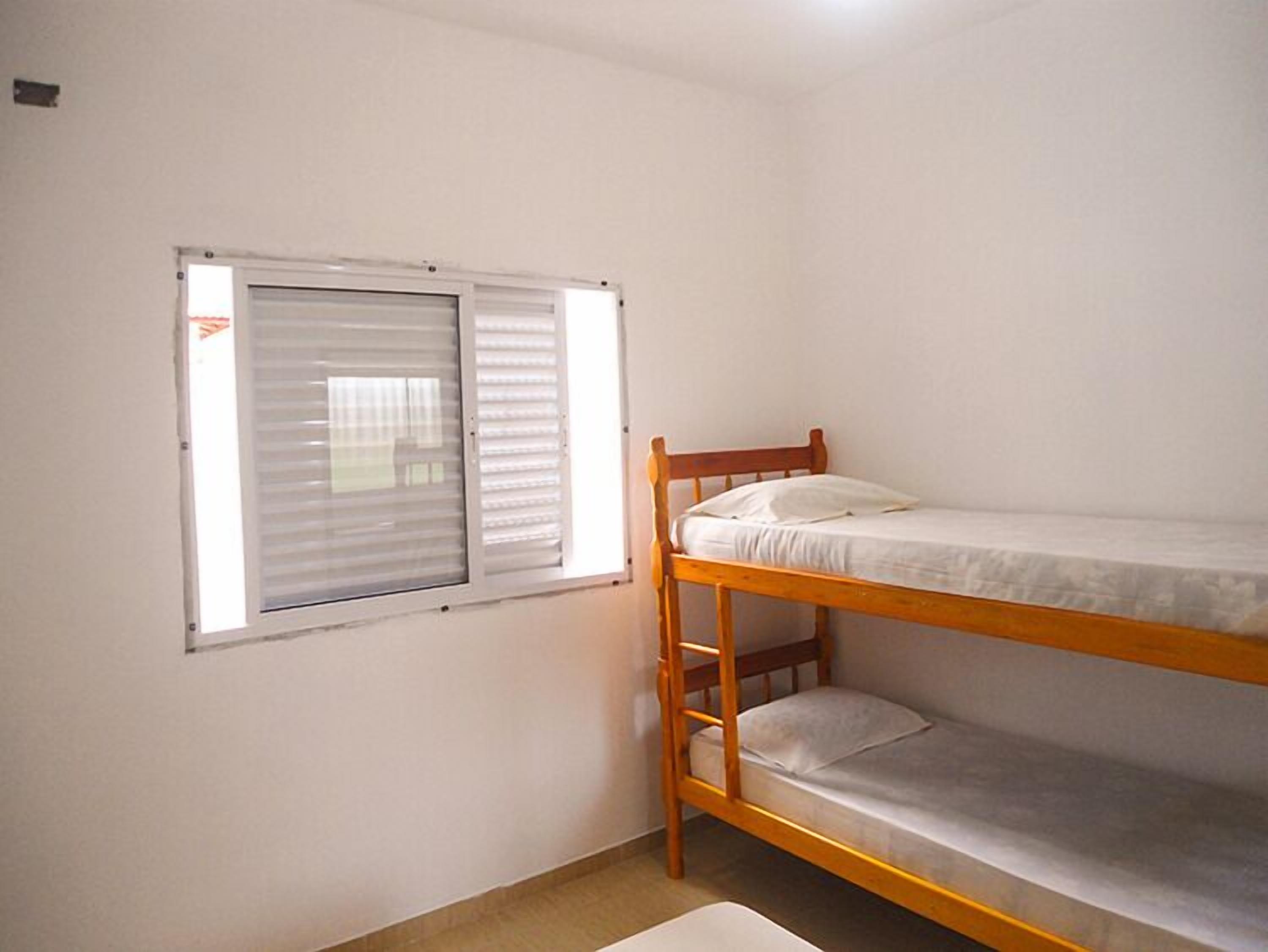 Een kleine kamer met een enkel bed en een houten stapelbed, voorzien van een raam met zonwering.