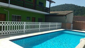 A swimming pool with a white lattice fence, a green building with a balcony, and a hilly landscape in the background. - Pousada Montanhas do Bem TeVi (Caraguatatuba)