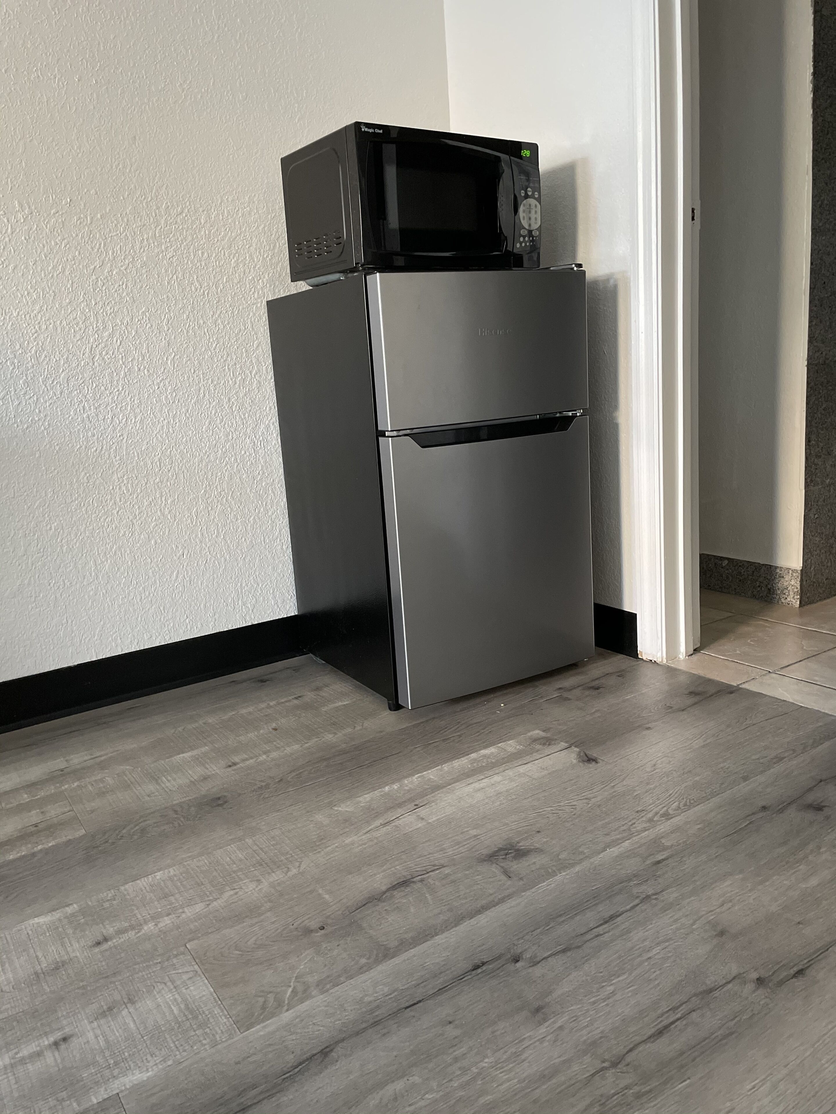 A compact kitchen with a stainless steel refrigerator and microwave, a wooden floor, and a white textured wall.
