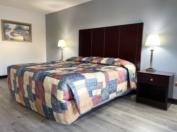 A neatly made bed with a colorful quilt, a dark headboard, a bedside table with a lamp, and a framed picture on the wall.