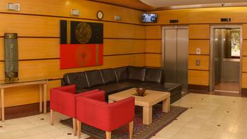 Un hall con un sofá de cuero, sillas rojas, una mesa de centro, un reloj, un televisor y un ascensor.