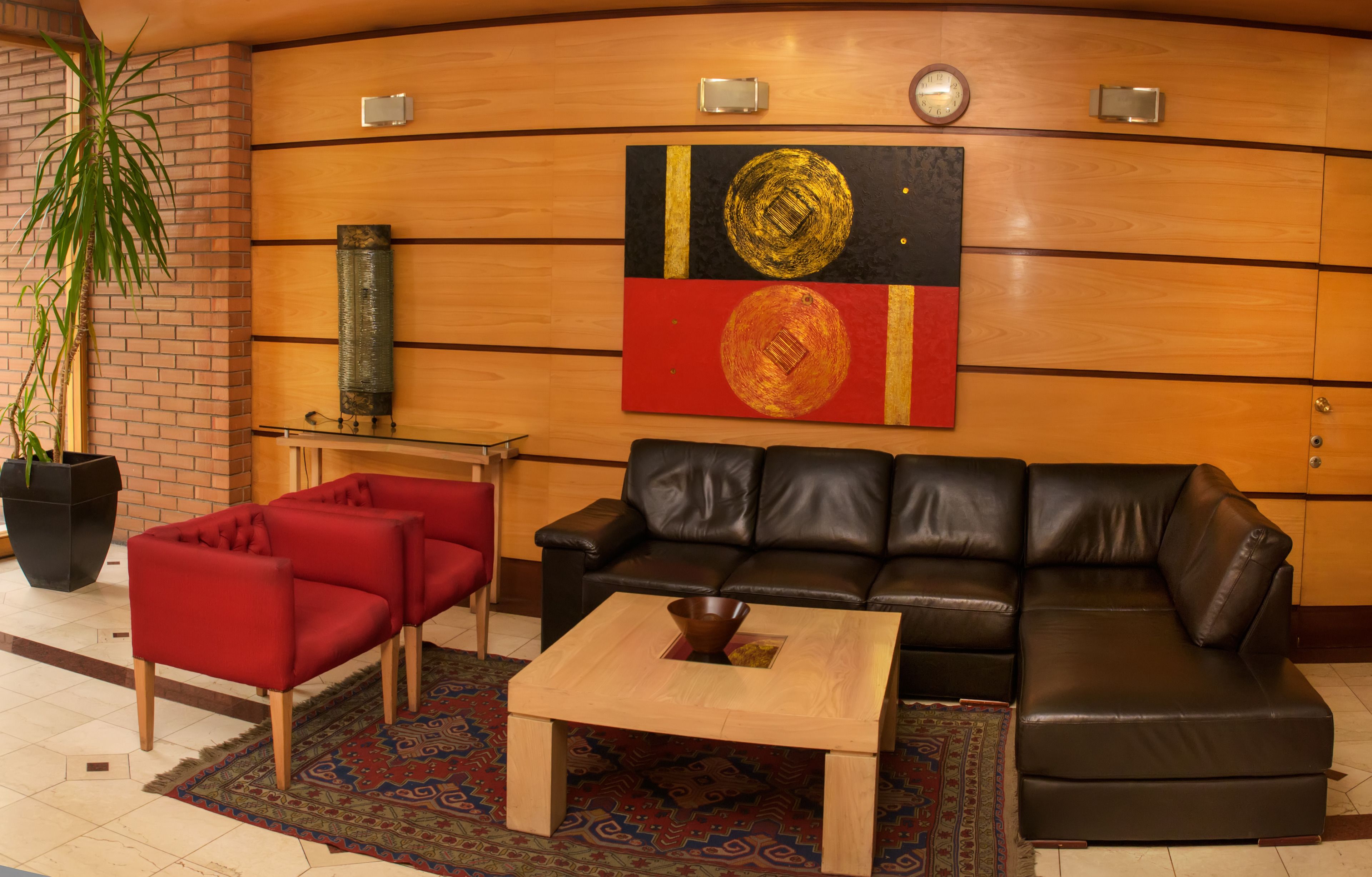 A lobby with a black leather sofa, red armchairs, a wooden coffee table, and a large abstract painting on the wall.