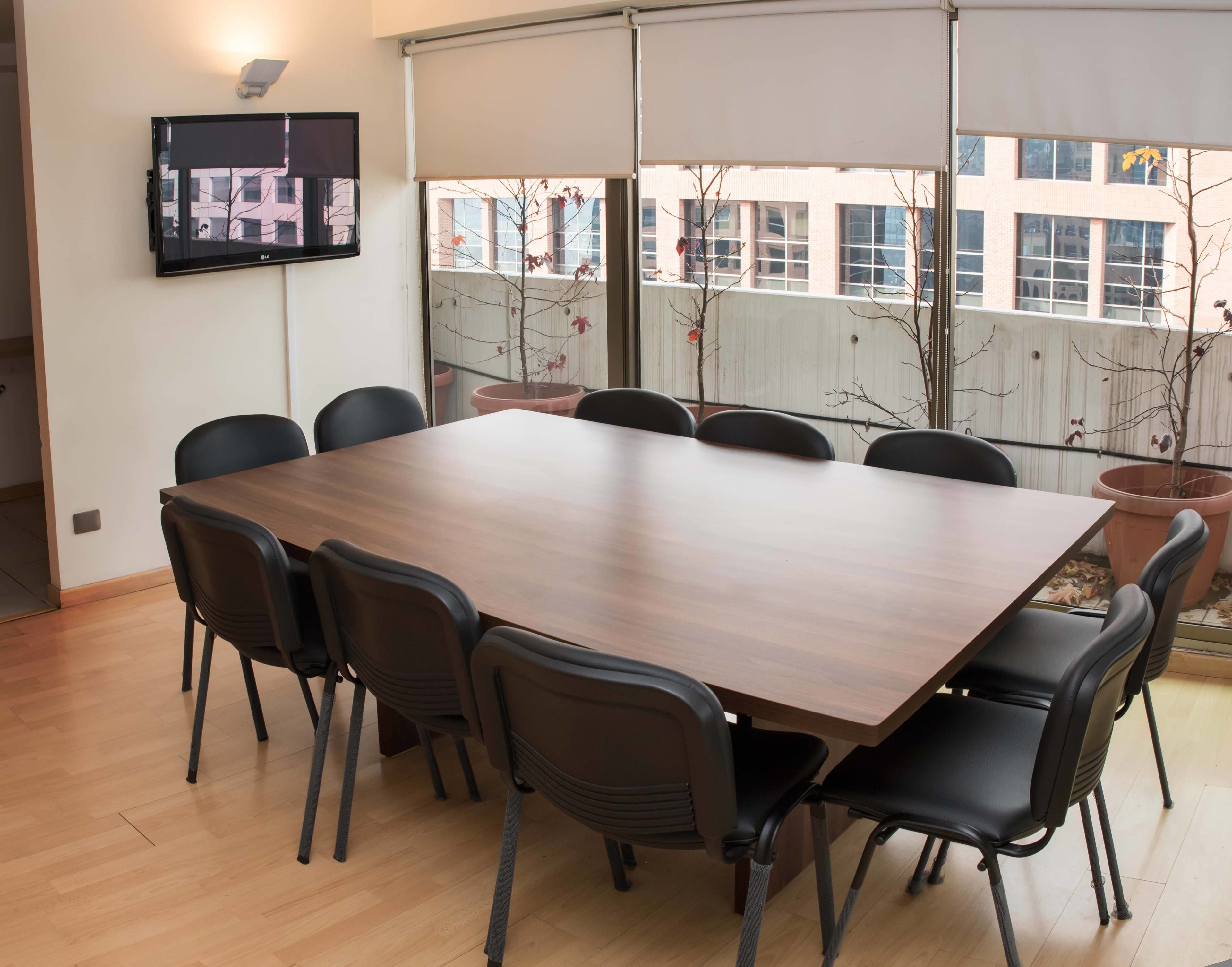 A conference room with a large wooden table, black chairs, a flat-screen TV on the wall, and a view of buildings through large windows.