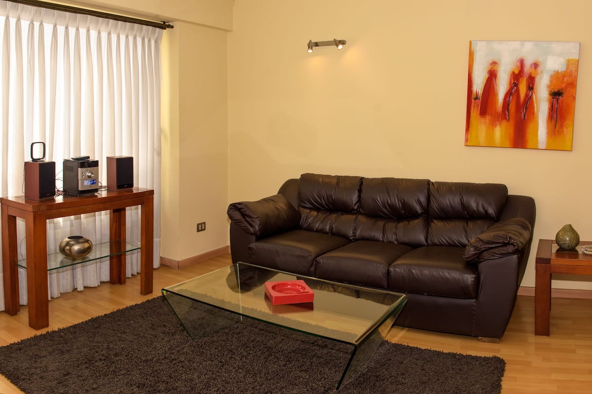 A living room with a brown leather sofa, a glass coffee table, a wooden side table, and a painting on the wall.