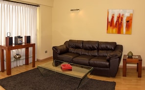 A living room with a brown leather sofa, a glass coffee table, a wooden side table, and a painting on the wall. - El Bosque Suites (Santiago)