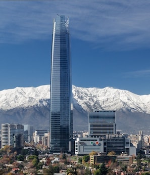 A tall skyscraper with a modern glass facade, surrounded by other buildings and a snow-capped mountain in the background. - El Bosque Suites (Santiago)