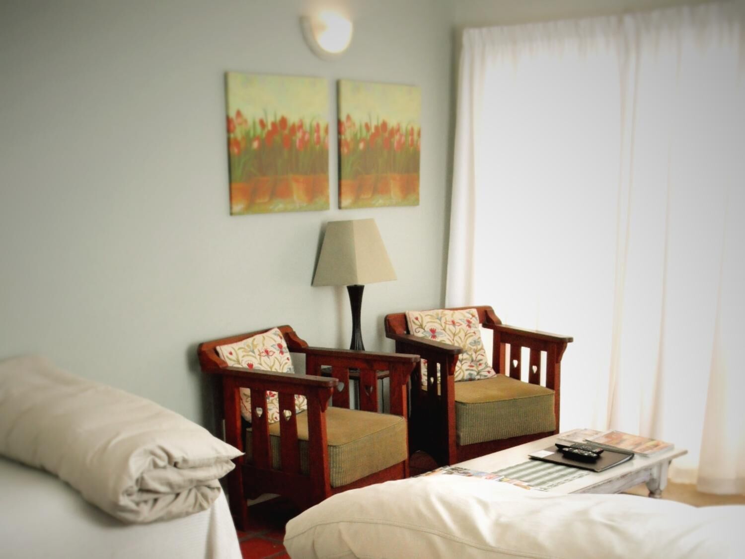 A hotel room with two wooden chairs, a bed, a nightstand, and two wall-mounted paintings.
