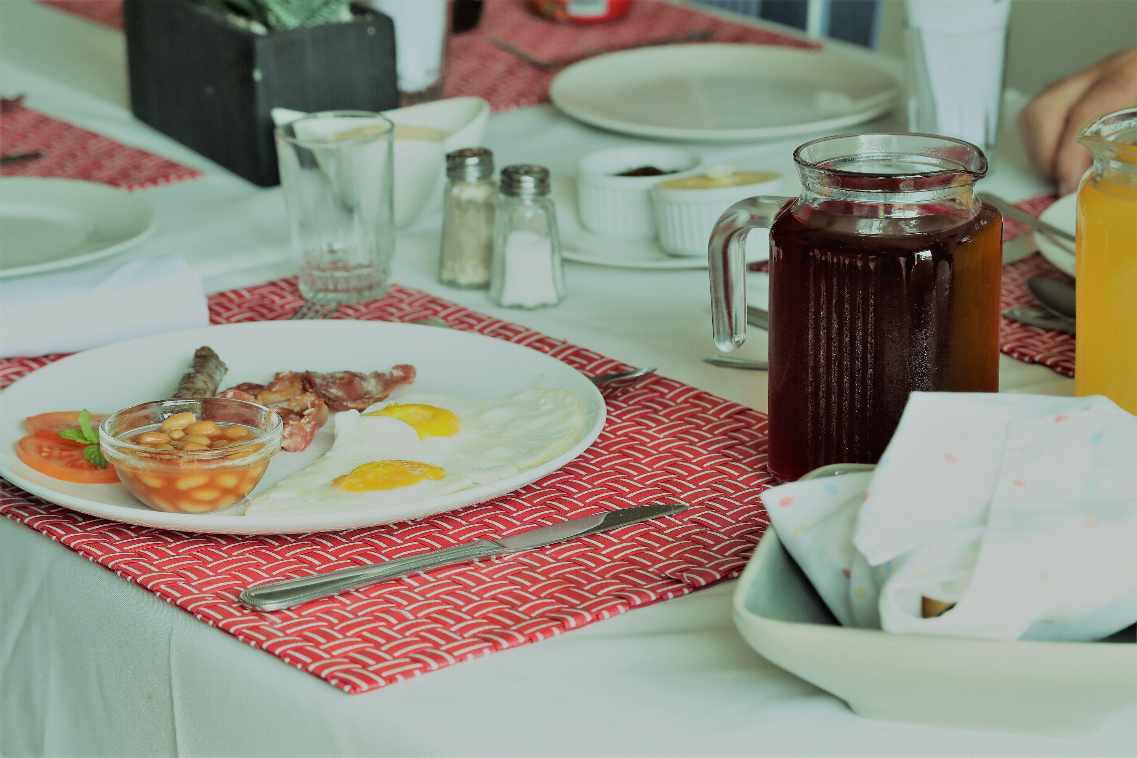 Une table mise pour le petit-déjeuner avec une assiette contenant des œufs, des haricots et des tomates, un pichet de jus, ainsi que des salières et des poivrières, le tout posé sur une table recouverte d’une nappe rouge à motifs.