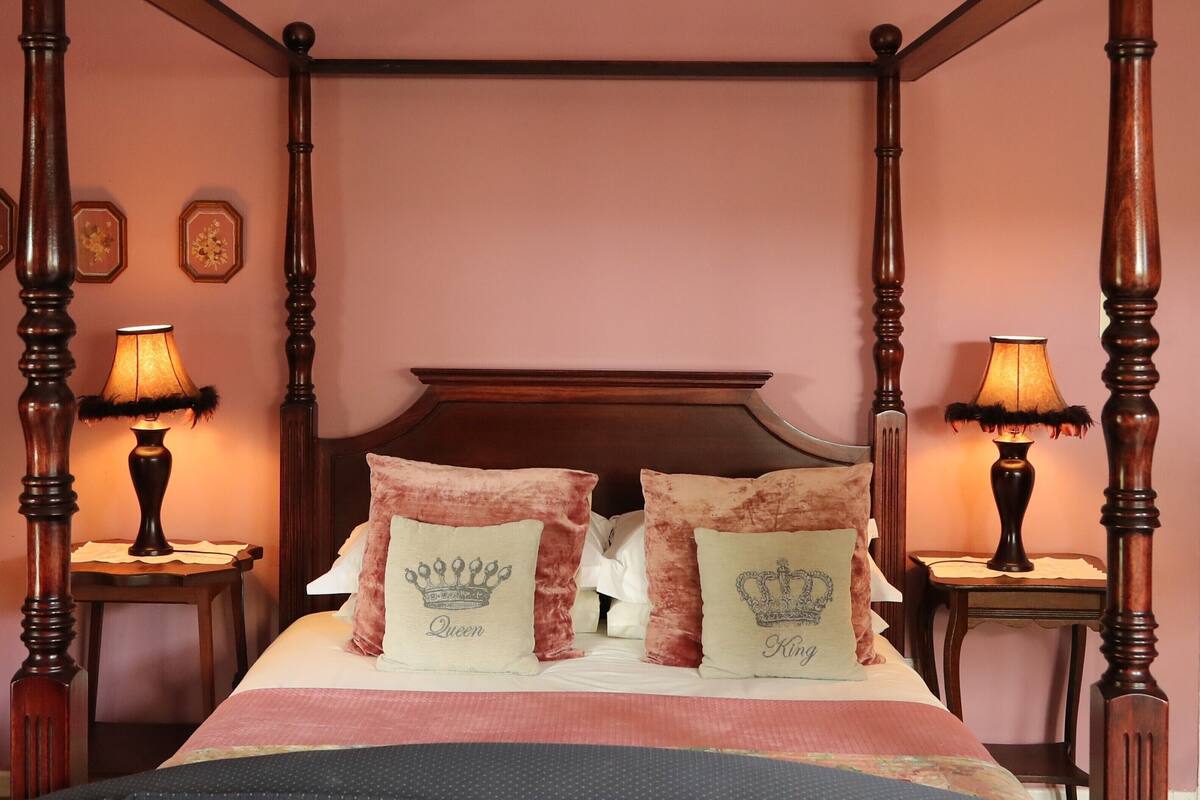 A four-poster bed with a wooden headboard, two decorative pillows with crown designs, and two bedside lamps on either side.