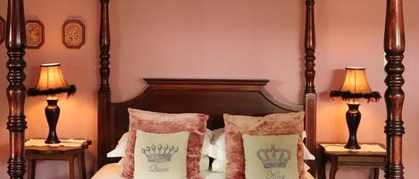 A four-poster bed with a wooden headboard, two decorative pillows with crown designs, and two bedside lamps on either side.