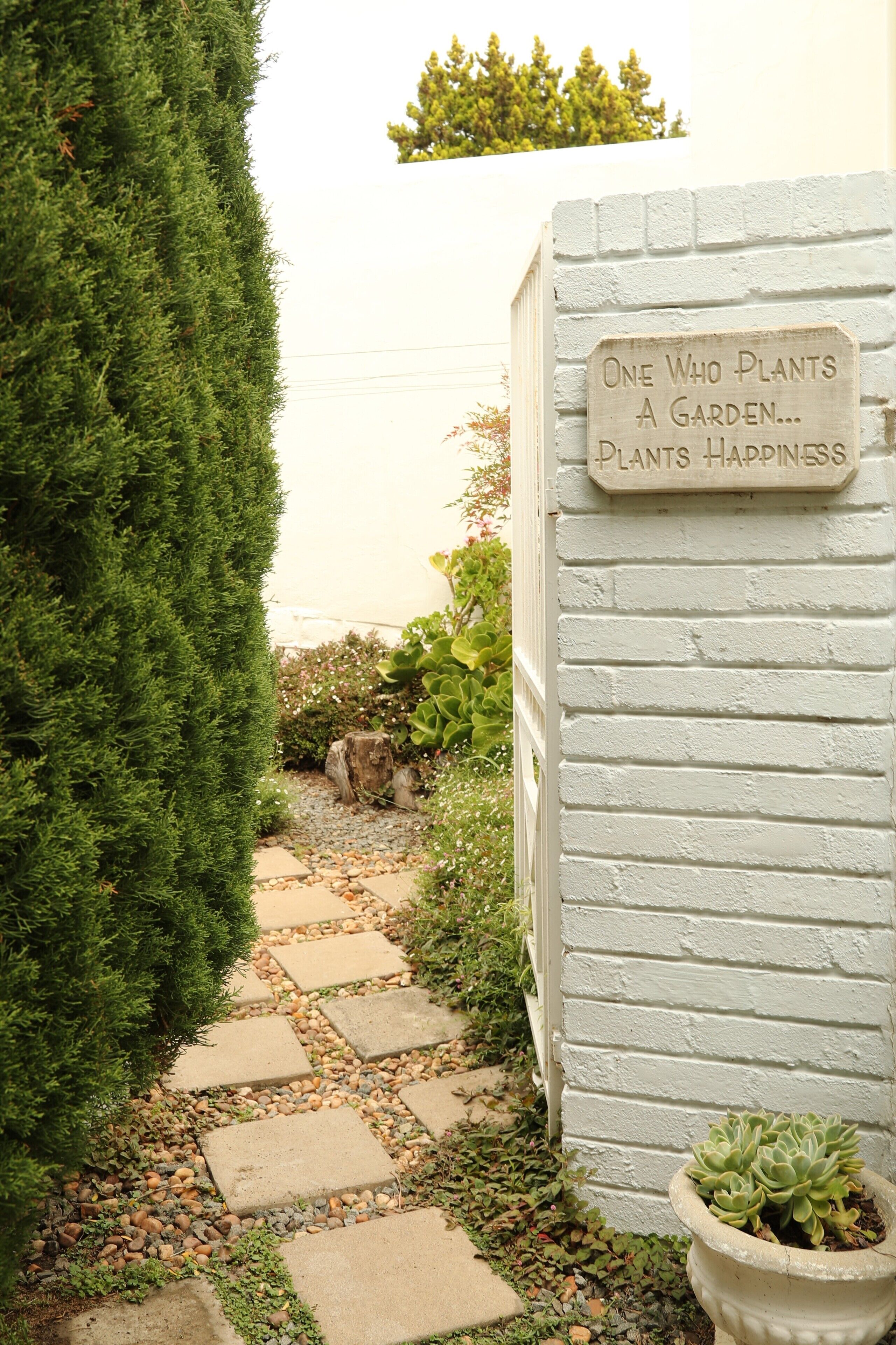 A garden pathway leading to a white brick wall with a sign that reads "One who plants a garden... plants happiness."