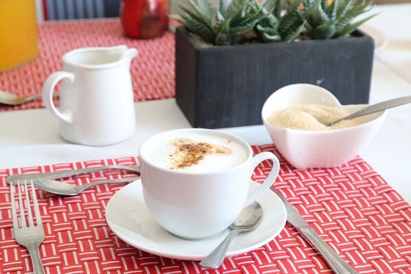 A white cup of coffee with a spoon on a white saucer, a white bowl with sugar, and a white pitcher on a red and white checkered tablecloth.