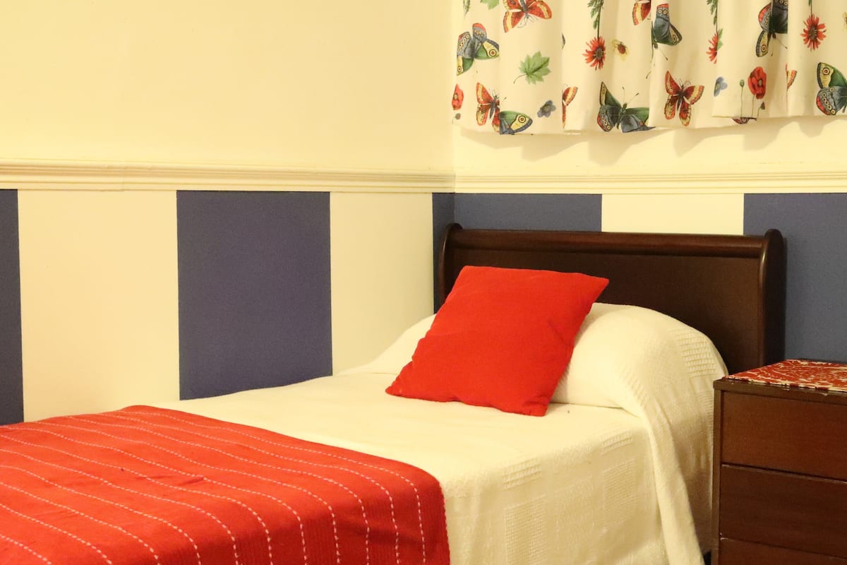 A neatly made bed with a red pillow and a red-striped blanket, a wooden nightstand with a patterned tablecloth, and a wall with a butterfly curtain.