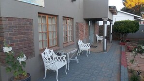 A building with a brick facade, two windows with wooden frames, and a covered outdoor seating area with white metal chairs and a table. - Aligeo Guest House (Senqu)