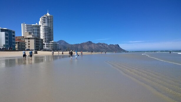 Ein Strand mit klarem Himmel, einem Berg im Hintergrund und mehreren Gebäuden entlang der Küste.