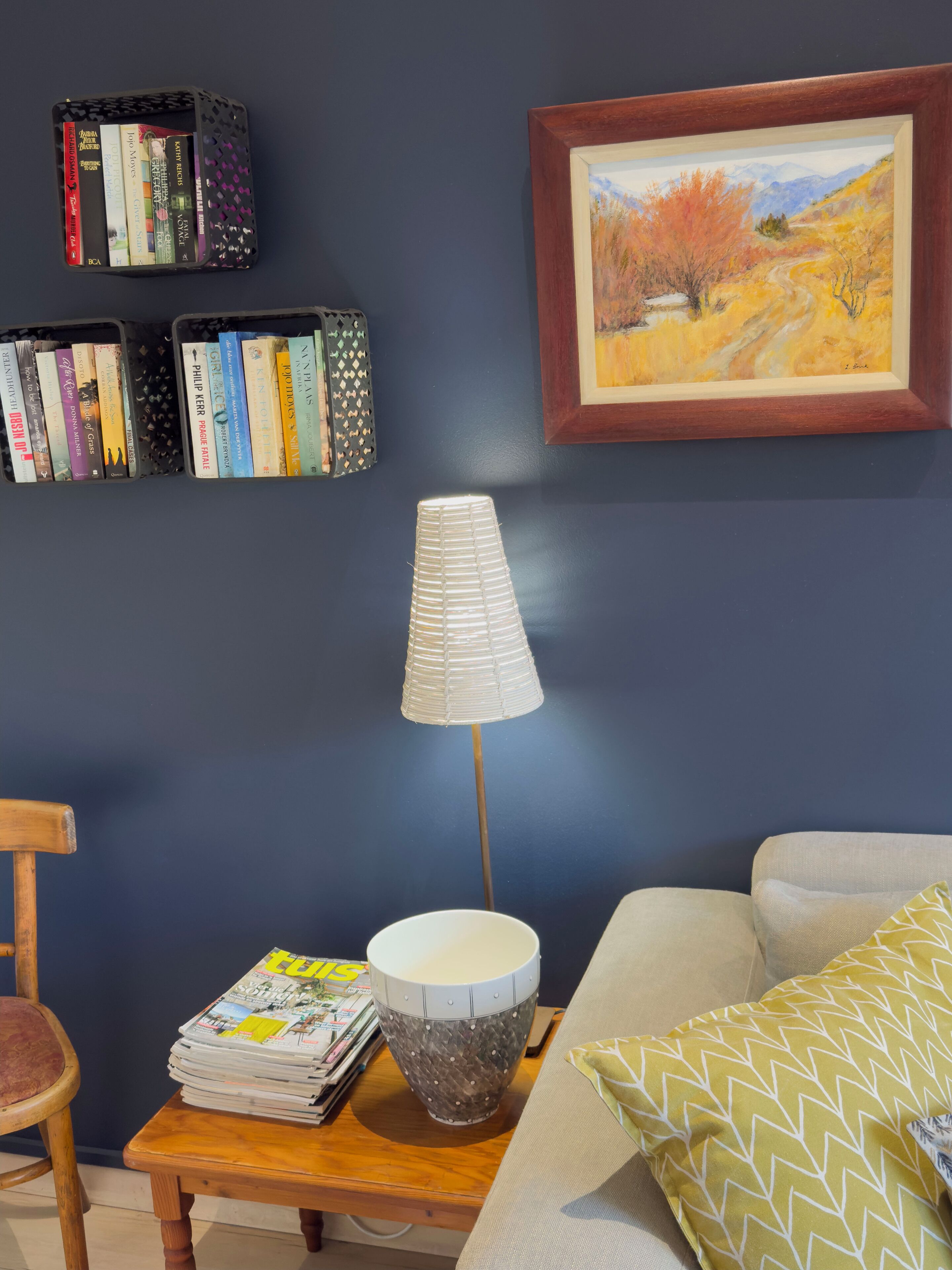 A room with a dark blue wall, a framed painting, a bookshelf, a lamp, a magazine, and a cushion.