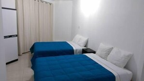 A room with two beds, each with a blue bedspread and white pillows. There is a window with blinds, a white door, and a small table with a lamp.