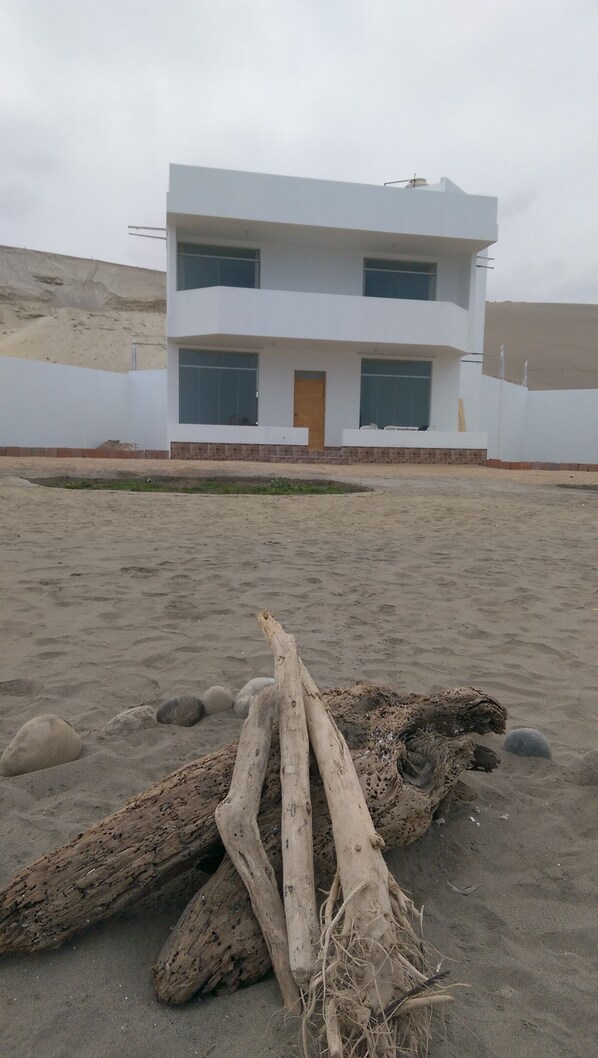 A modern two-story building with a flat roof and large windows. In front of the building, there is a sandy area with scattered rocks and driftwood. - Camana Beach House (Samuel Pastor)