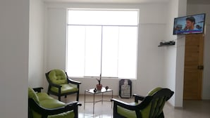 A living room with two green chairs, a small table with a vase, and a television on the wall.
