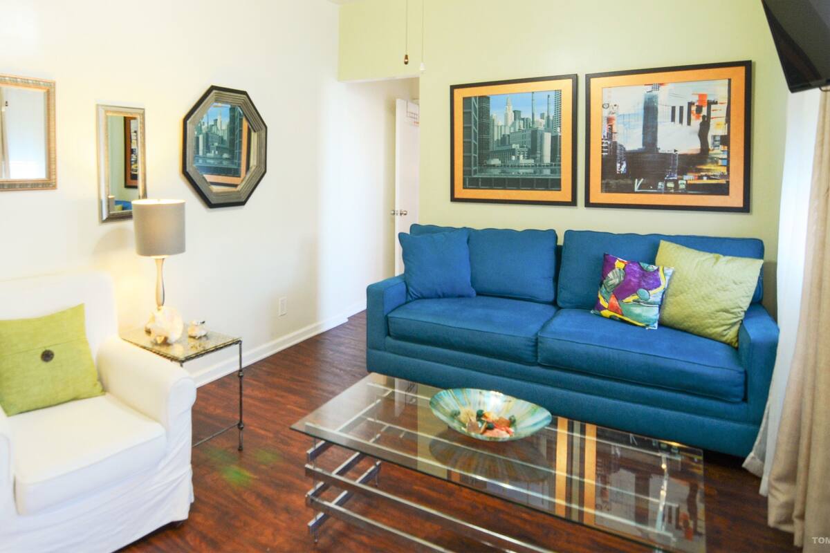 Una sala de estar con un sillón blanco, un sofá azul, una mesa de centro de vidrio y cuadros en las paredes.
