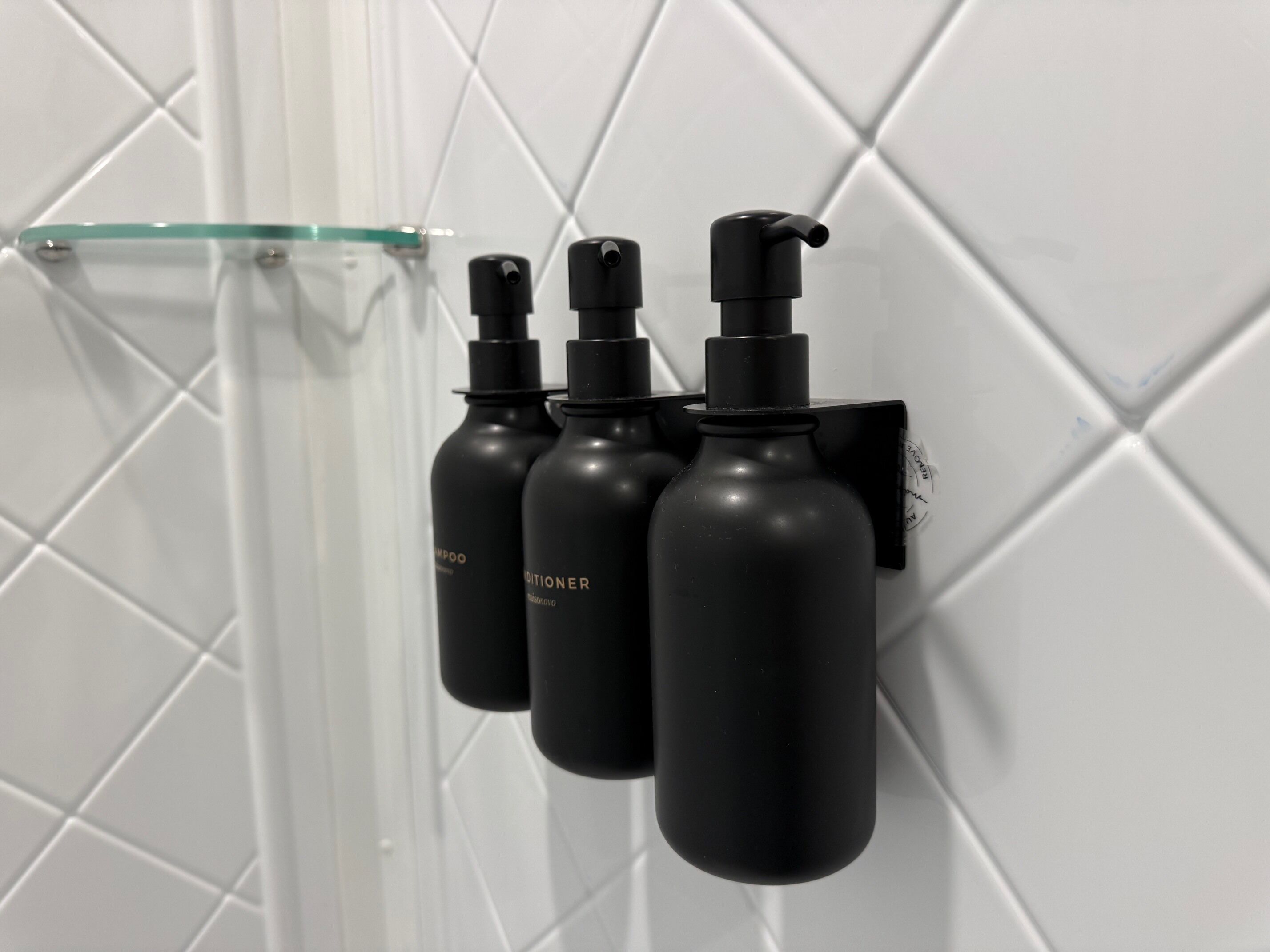 A bathroom with a white tiled wall and three black bottles of shampoo and conditioner mounted on the wall.