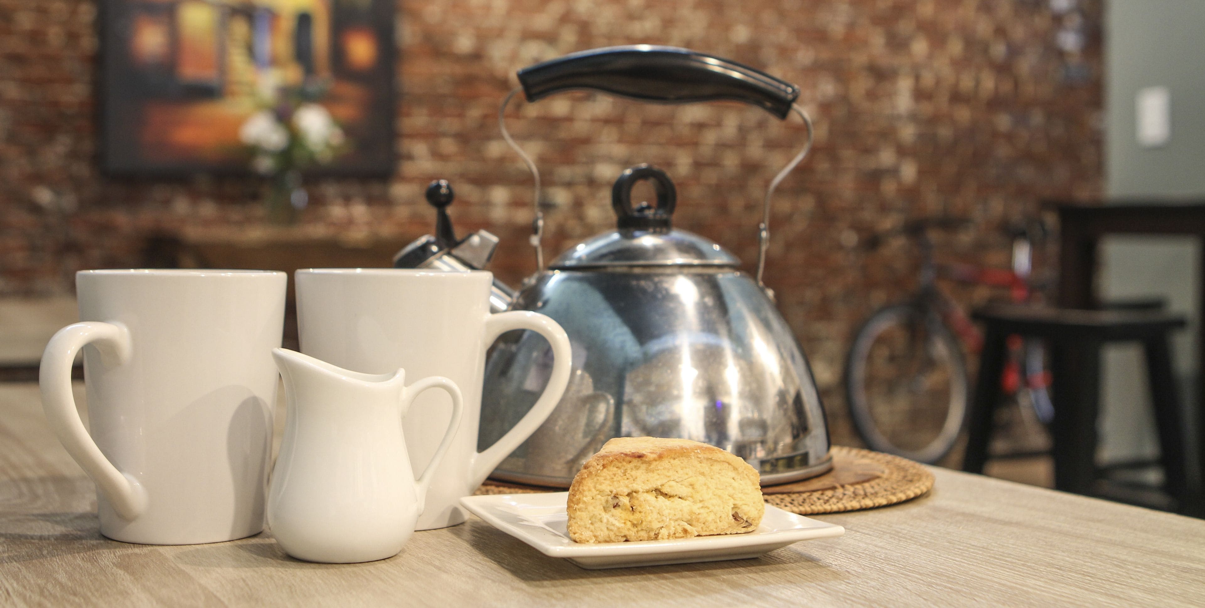 A kitchen area with a kettle, two white mugs, a white pitcher, and a plate with a biscuit.