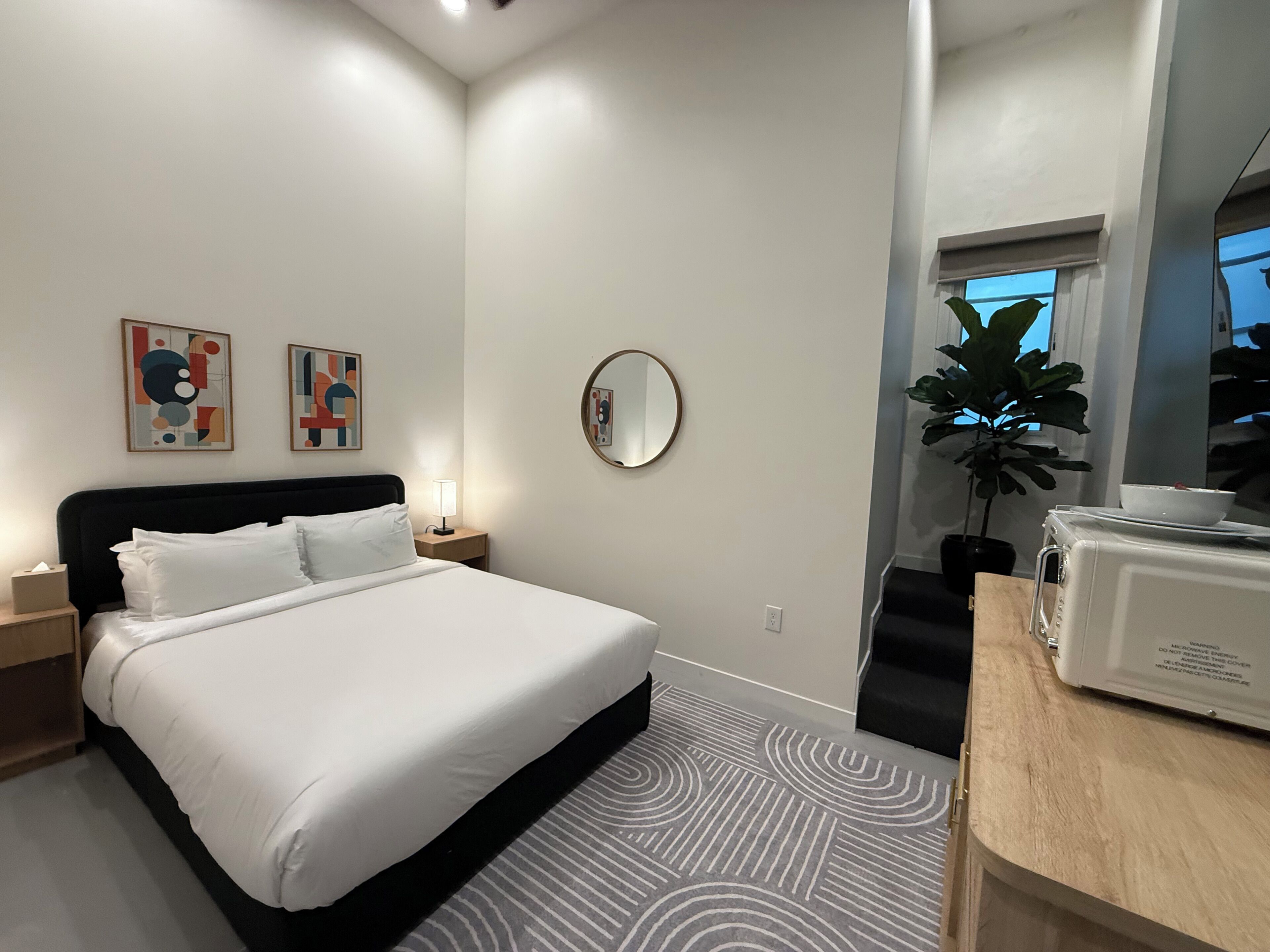Une chambre d’hôtel moderne équipée d’un lit, d’une table de chevet, d’une lampe, d’une œuvre d’art, d’un miroir, d’une plante en pot et d’un four à micro-ondes.