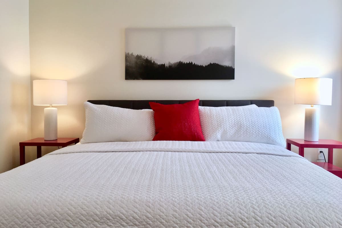 Una cama bien hecha con ropa blanca y una única almohada roja, flanqueada por dos mesitas de noche con lámparas, en una habitación con un cuadro de paisaje grande en la pared.