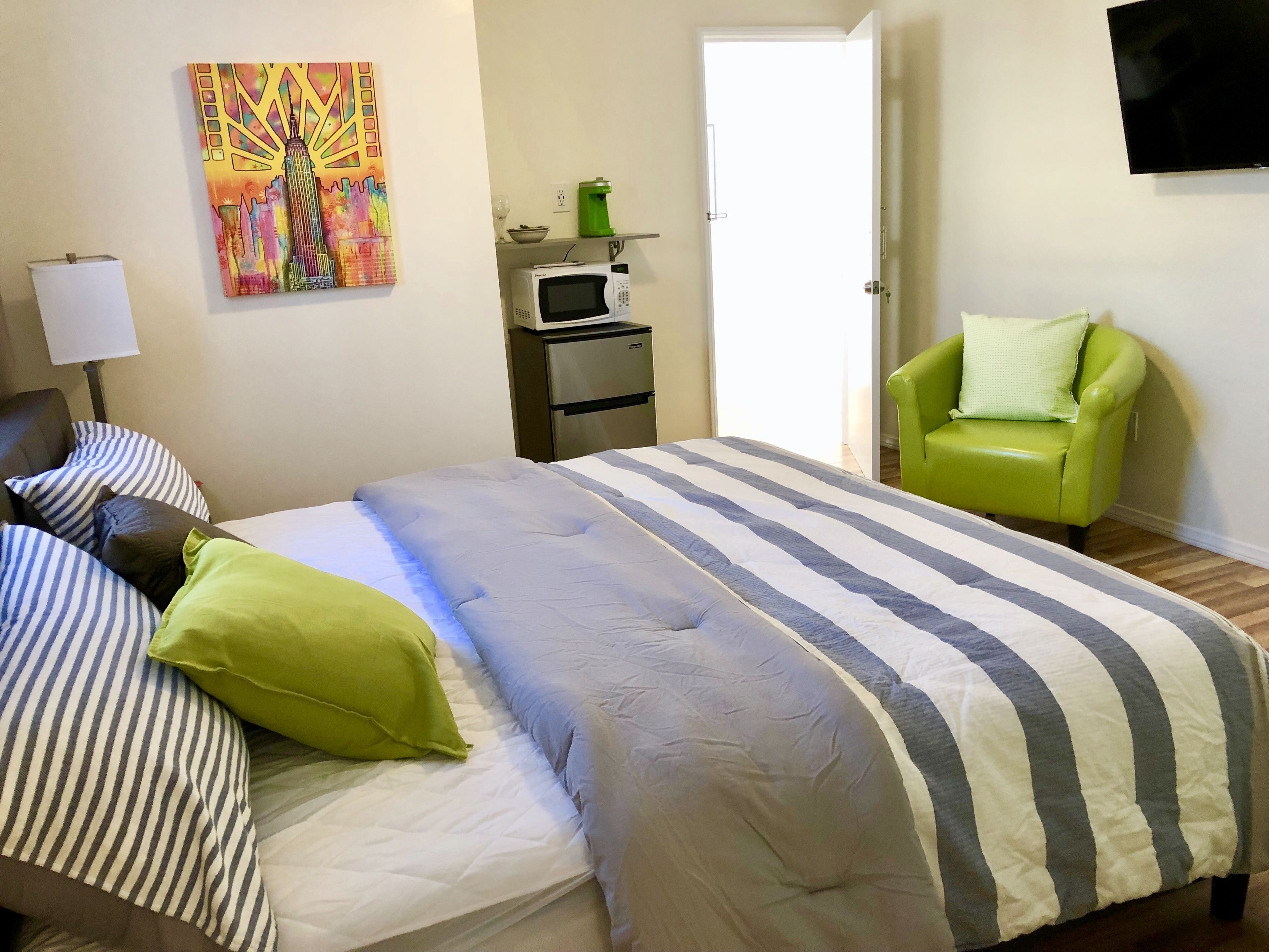 A hotel room with a bed, a green armchair, a microwave, a painting, and a TV on the wall.