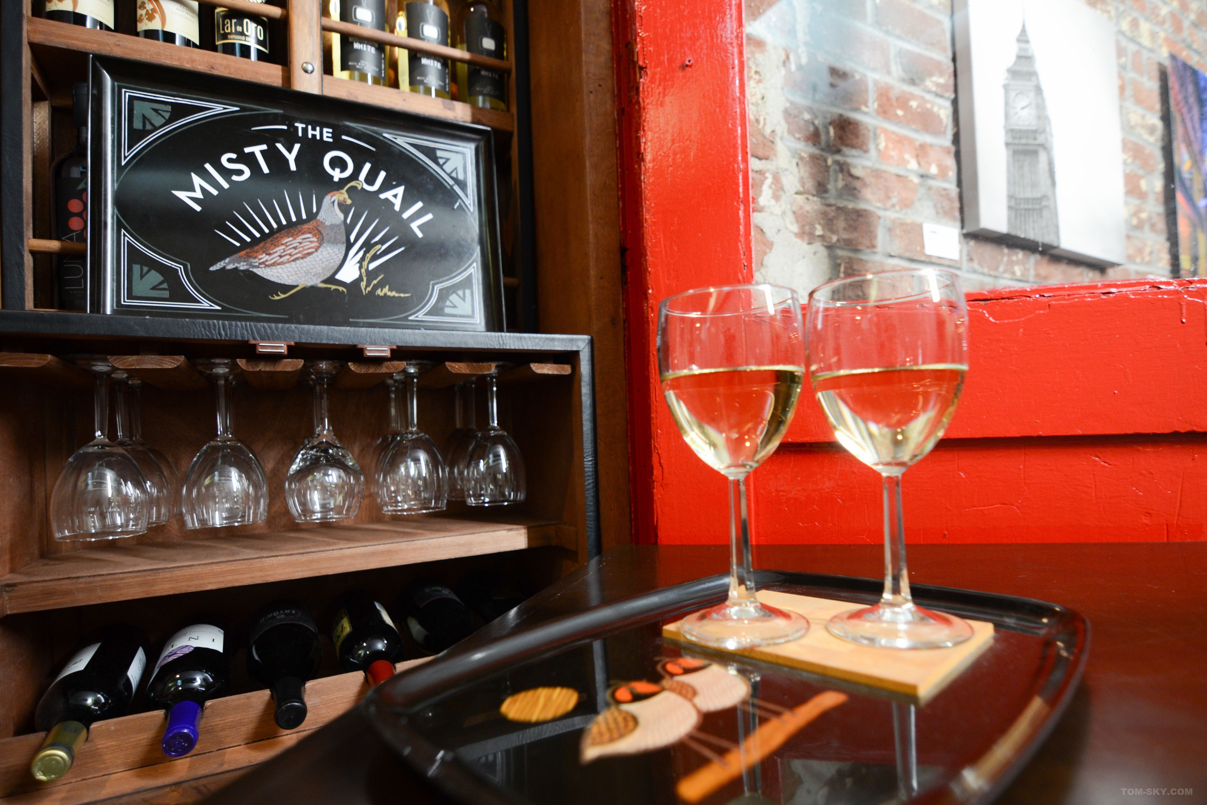 A bar area with a sign reading "The Misty Quail," wine bottles, and glasses on a tray.