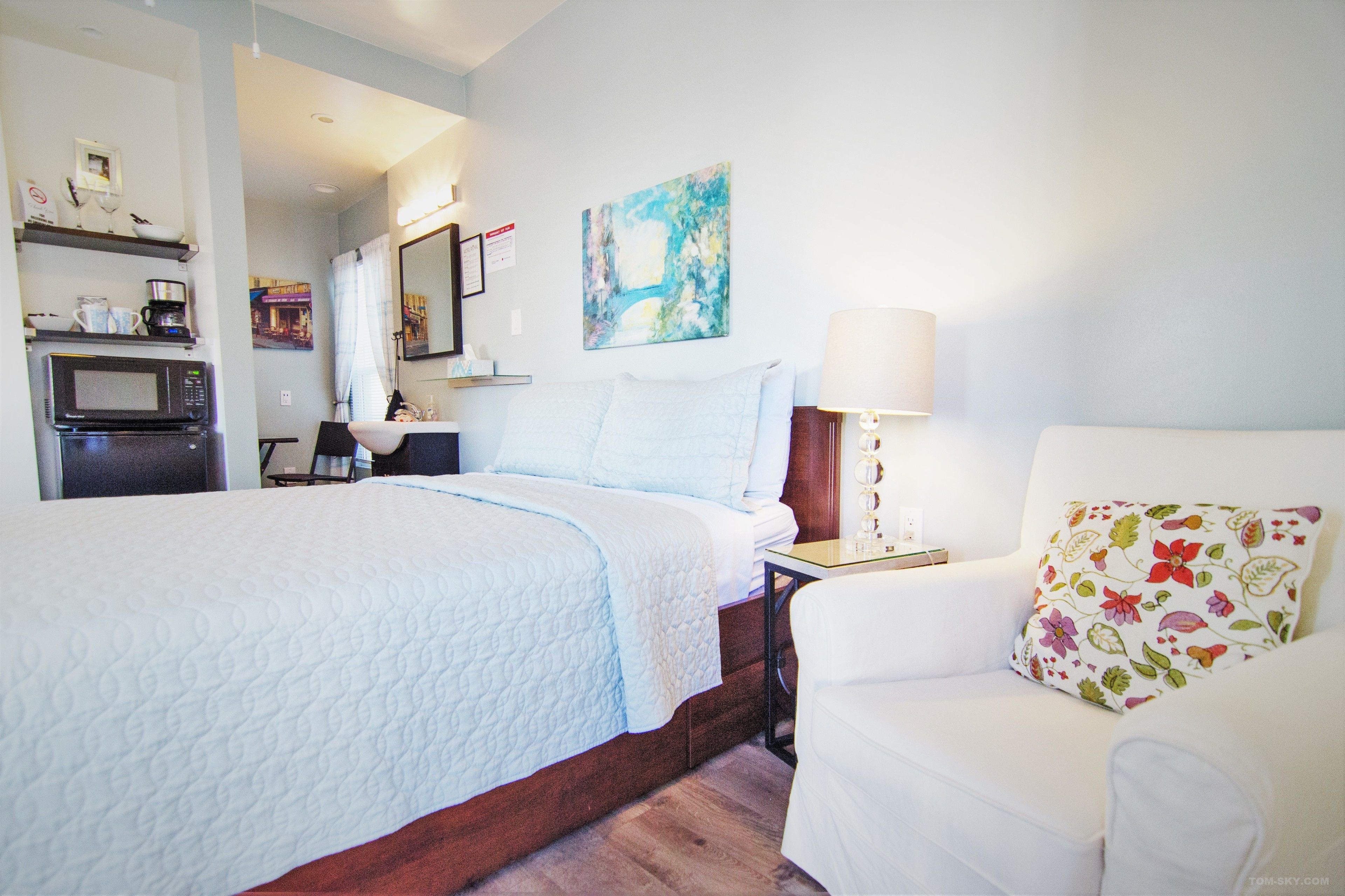 A hotel room with a bed, a white armchair, a lamp, a painting, a microwave, and a coffee maker.