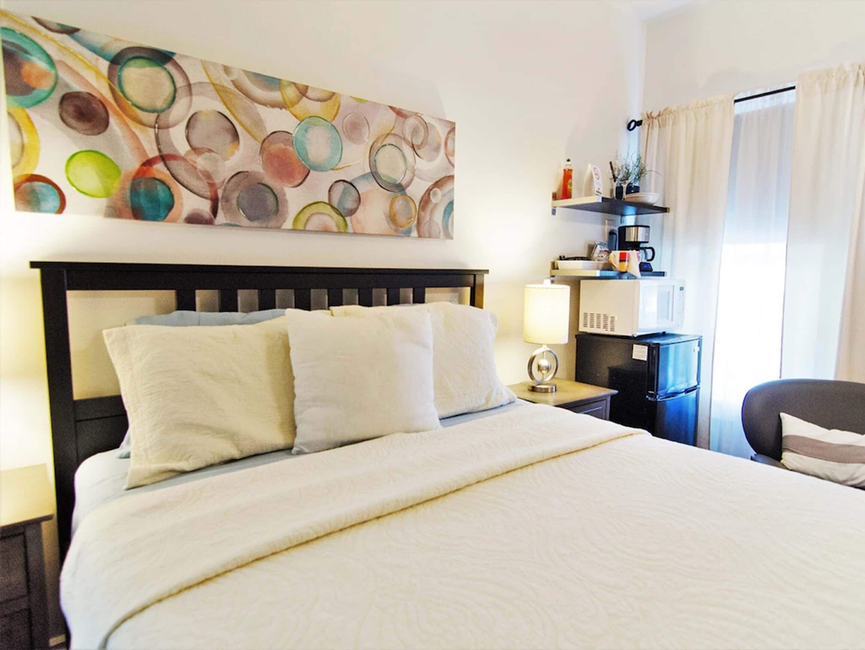 A hotel room with a large bed, a headboard, a bedside table with a lamp, a small shelf with toiletries, and a window with curtains.