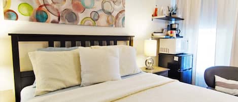 A hotel room with a large bed, a headboard, a bedside table with a lamp, a small shelf with toiletries, and a window with curtains.