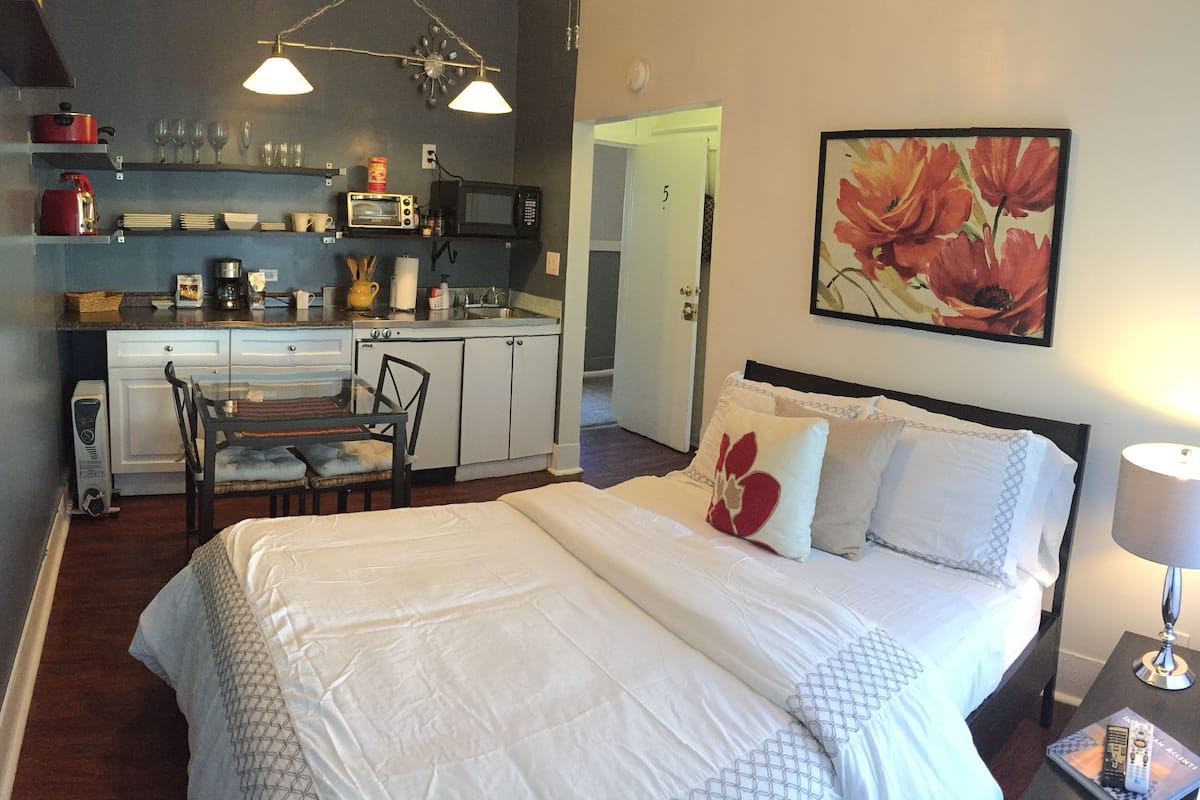 Una habitación de hotel compacta con kitchenette, una cama con ropa blanca, una mesita de noche con lámpara y una obra de arte floral en la pared.
