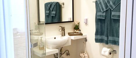 A bathroom with a glass shower enclosure, a white sink with a mirror above it, a toilet, and a blue rug on the floor.