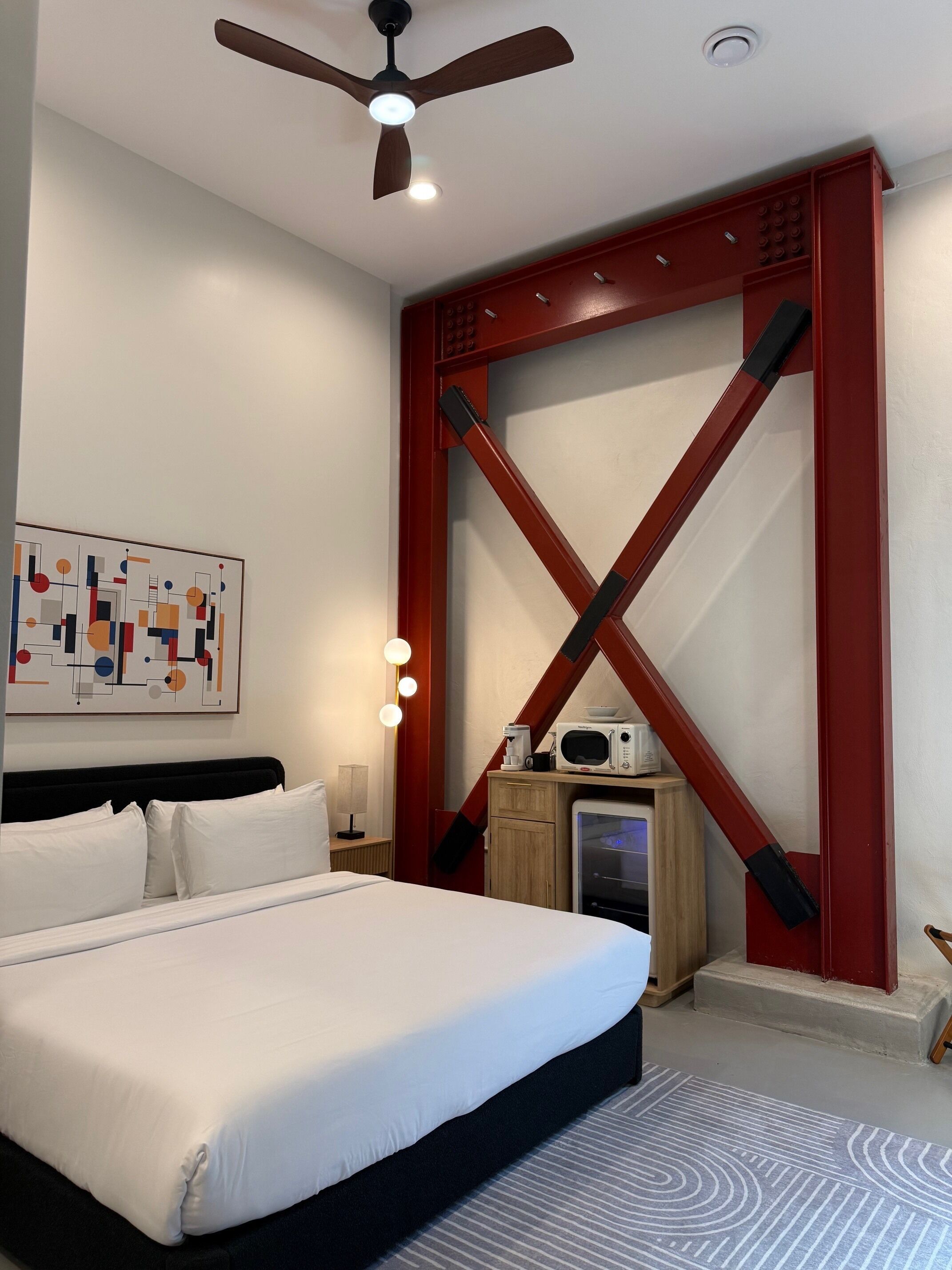 Une chambre d’hôtel comprenant un lit, un ventilateur de plafond, une œuvre d’art accrochée au mur, une table de chevet avec une lampe et un four à micro-ondes posé sur un meuble en bois.