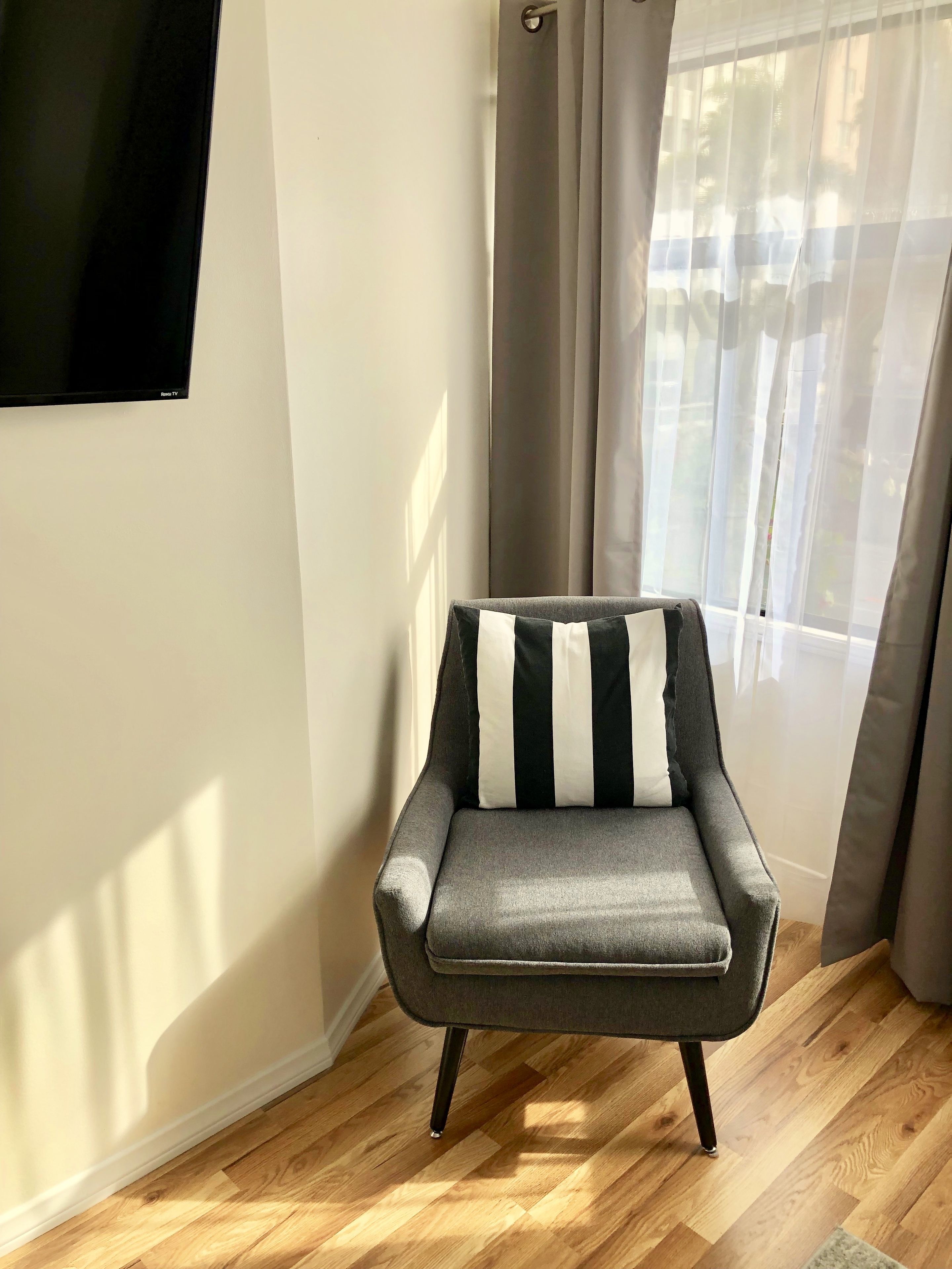 A corner room with a grey armchair, a flat-screen TV, and a window with sheer curtains.