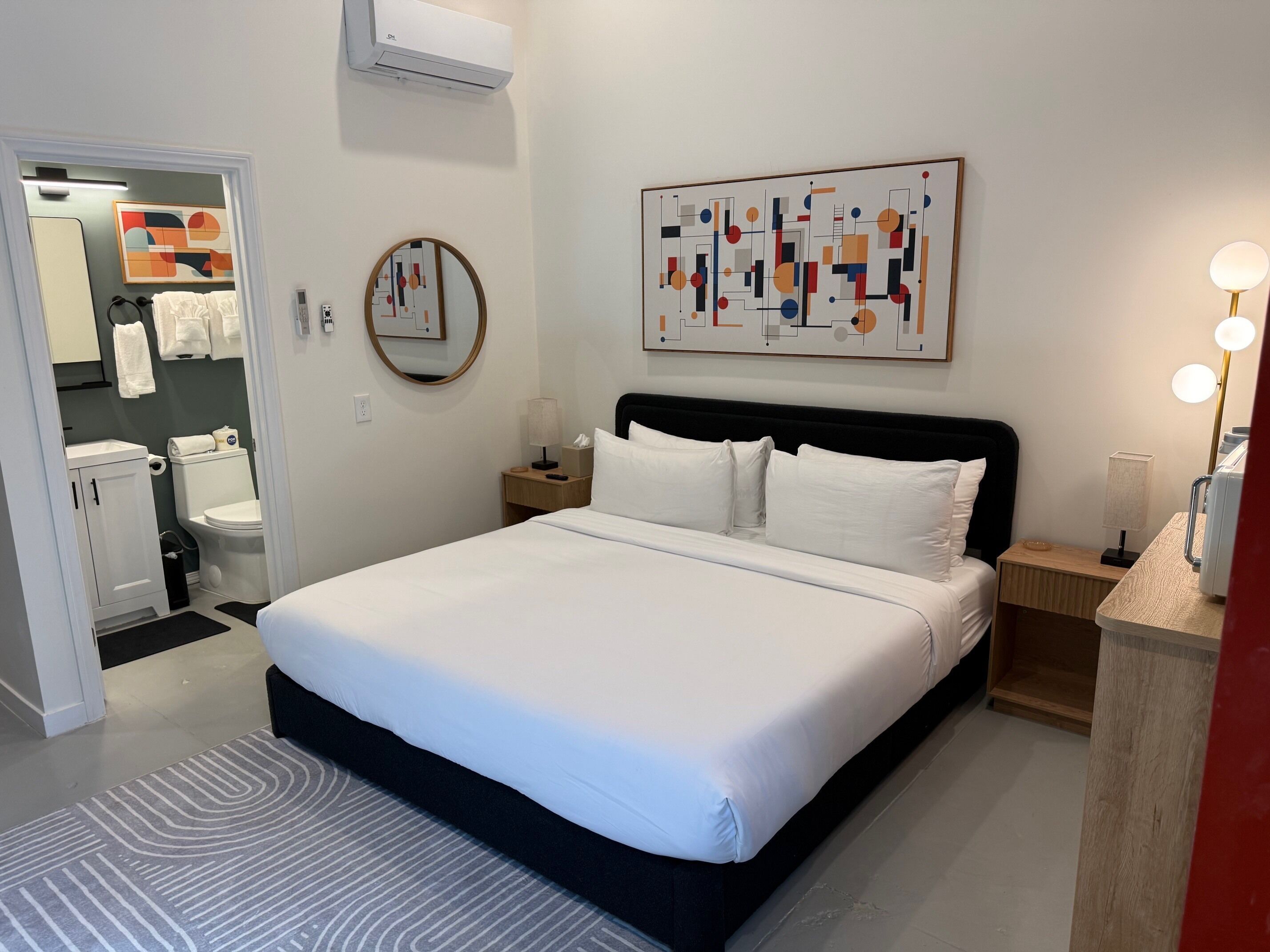 A hotel room with a large bed, a bedside table with a lamp, a wall-mounted air conditioner, a mirror, and a bathroom visible through an open door.