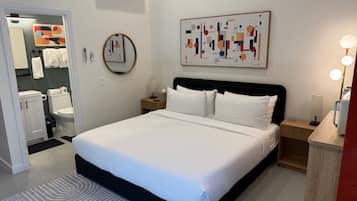 A hotel room with a large bed, a bedside table with a lamp, a wall-mounted air conditioner, a mirror, and a bathroom visible through an open door.