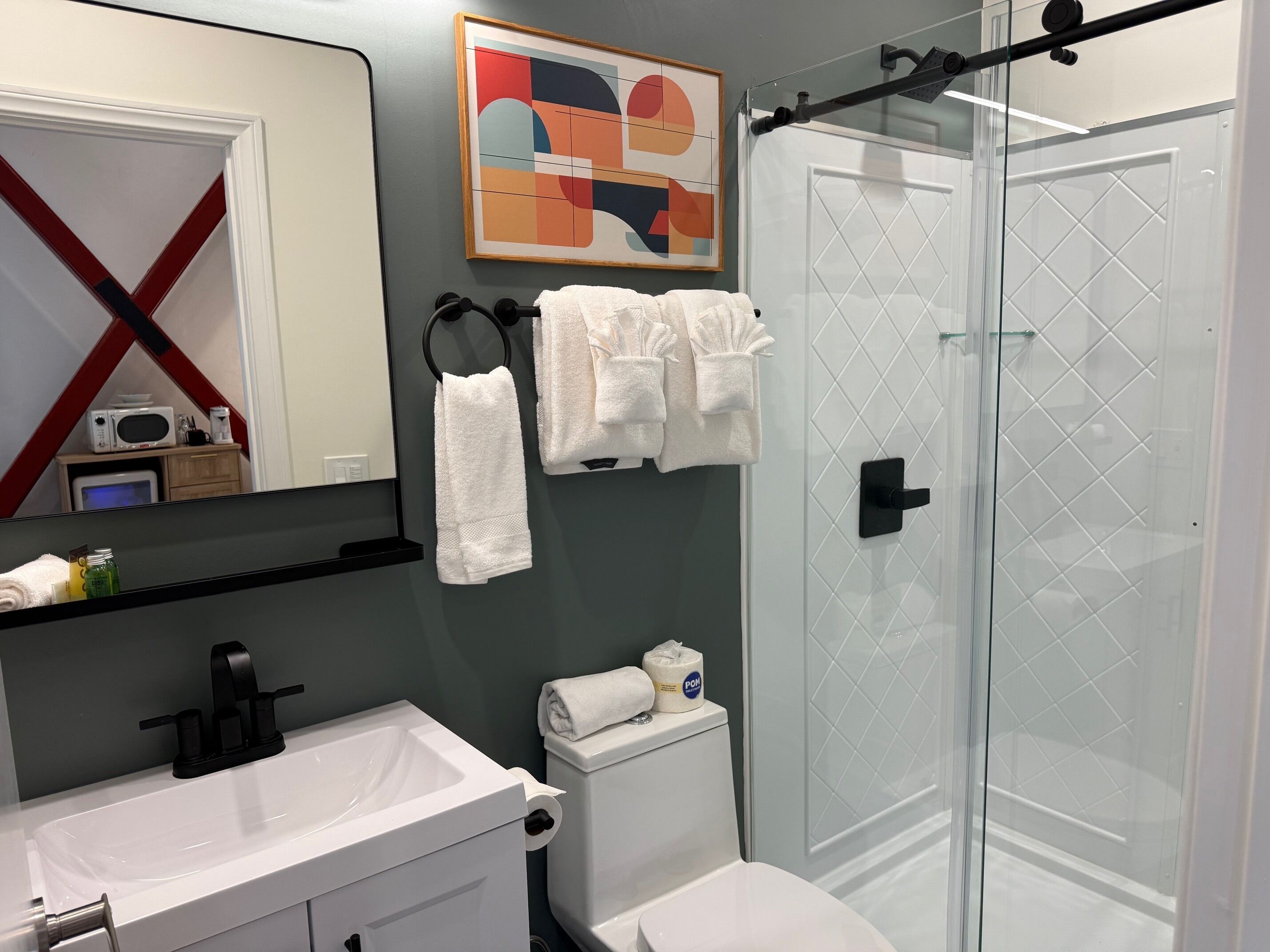 A bathroom with a white sink, toilet, and glass shower enclosure. There is a towel rack with towels and a framed abstract painting above the mirror.