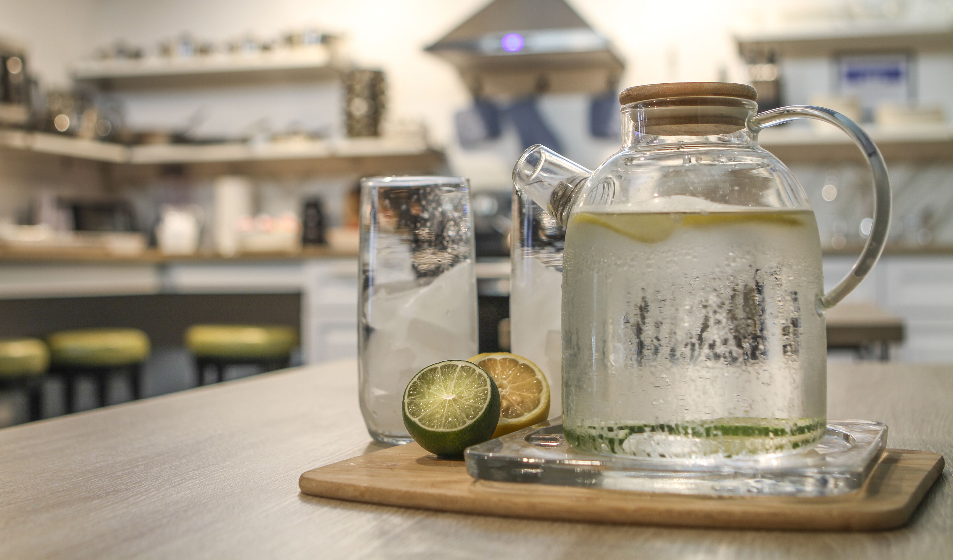 A clear glass pitcher with a wooden handle and a wooden lid, placed on a wooden board. A glass of water and a half-cut lime are also visible on the board.