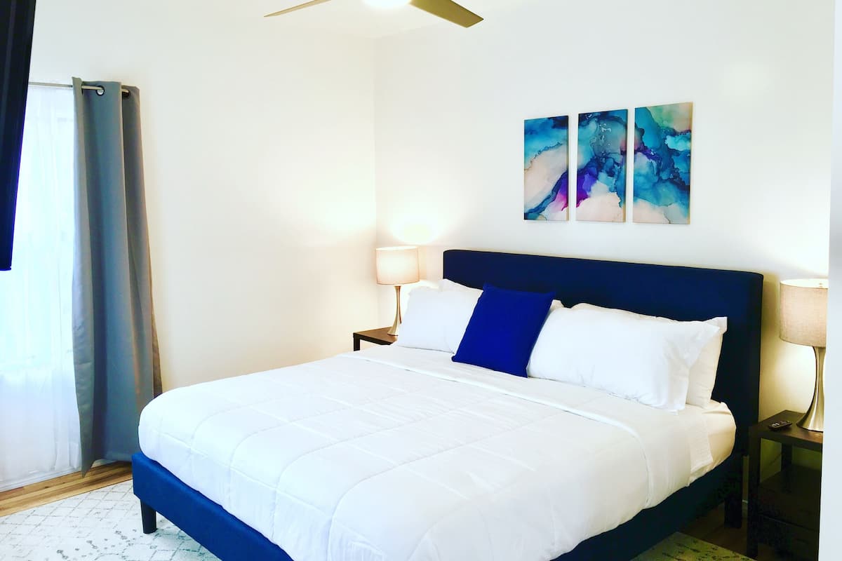Una habitación de hotel con una cama, ventilador de techo, dos cuadros en la pared y una mesita de noche con lámpara.