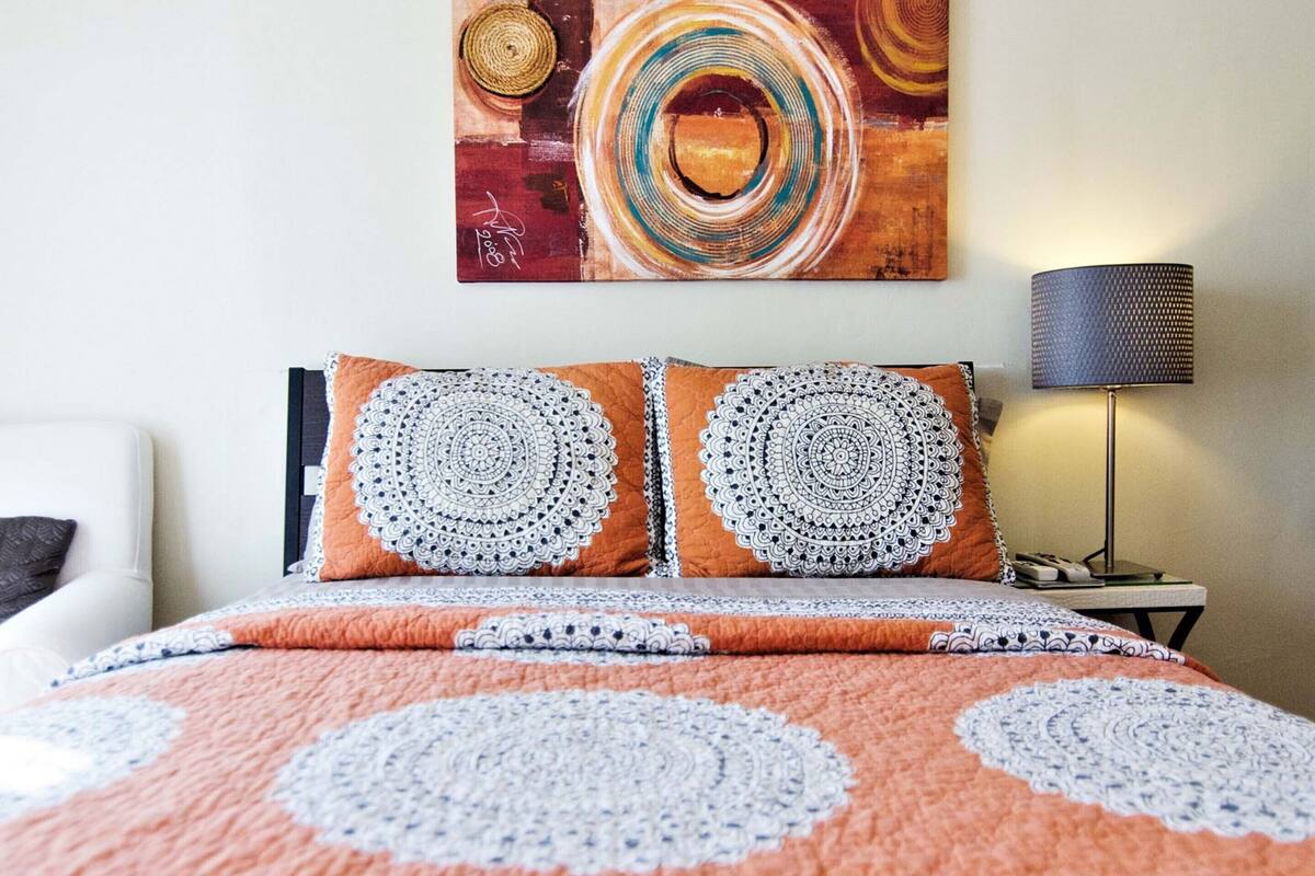 Una cama con un edredón estampado, dos almohadas con diseños circulares, un cuadro abstracto colgado en la pared y una lámpara de noche.