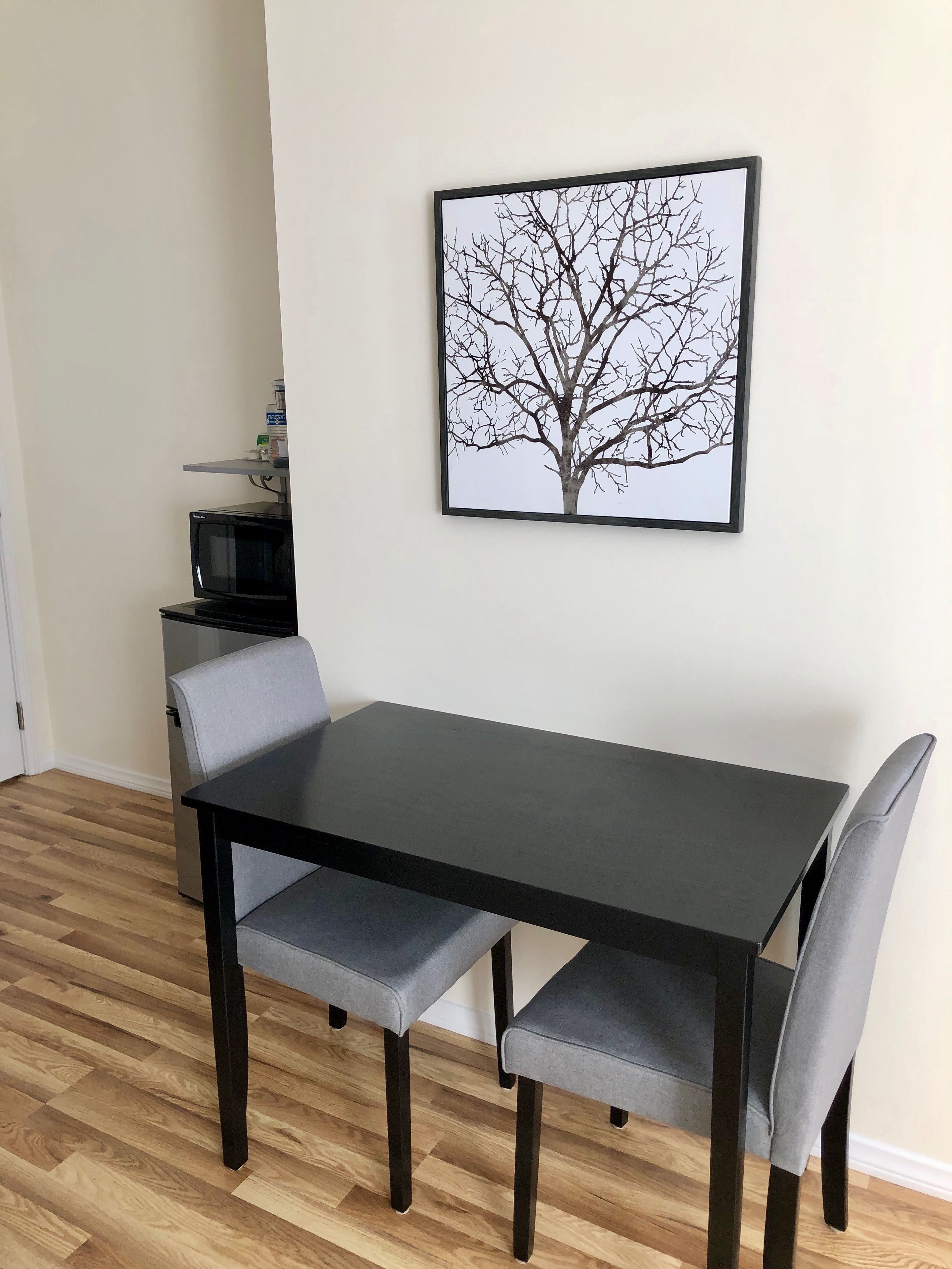 A dining area with a black table and grey chairs, a framed picture of a tree on the wall, and a microwave on a shelf.