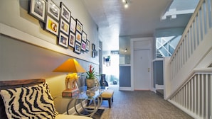 A hallway with a staircase, a zebra-patterned pillow on a sofa, a glass coffee table with decorative items, and framed pictures on the wall. - Hotel Royal (Long Beach)