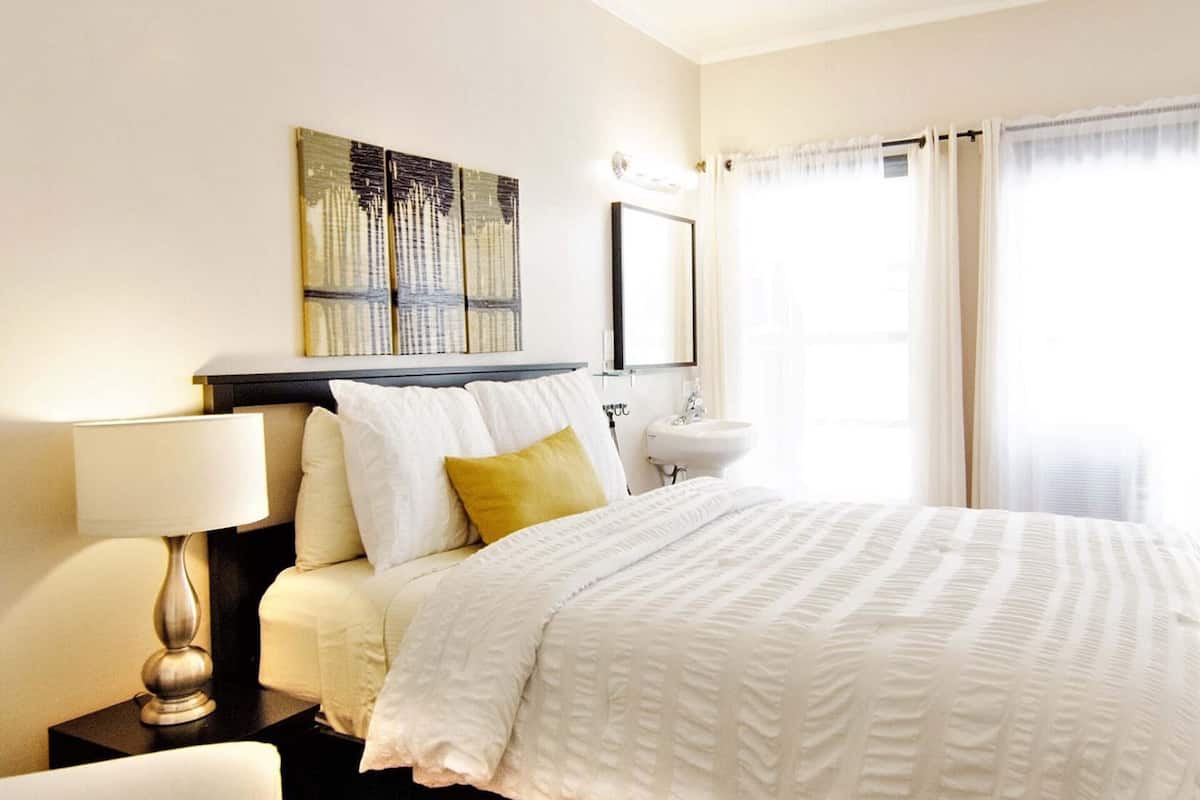 Habitación de hotel ordenada con cama, mesita de noche con lámpara, espejo en la pared y ventana con cortinas.